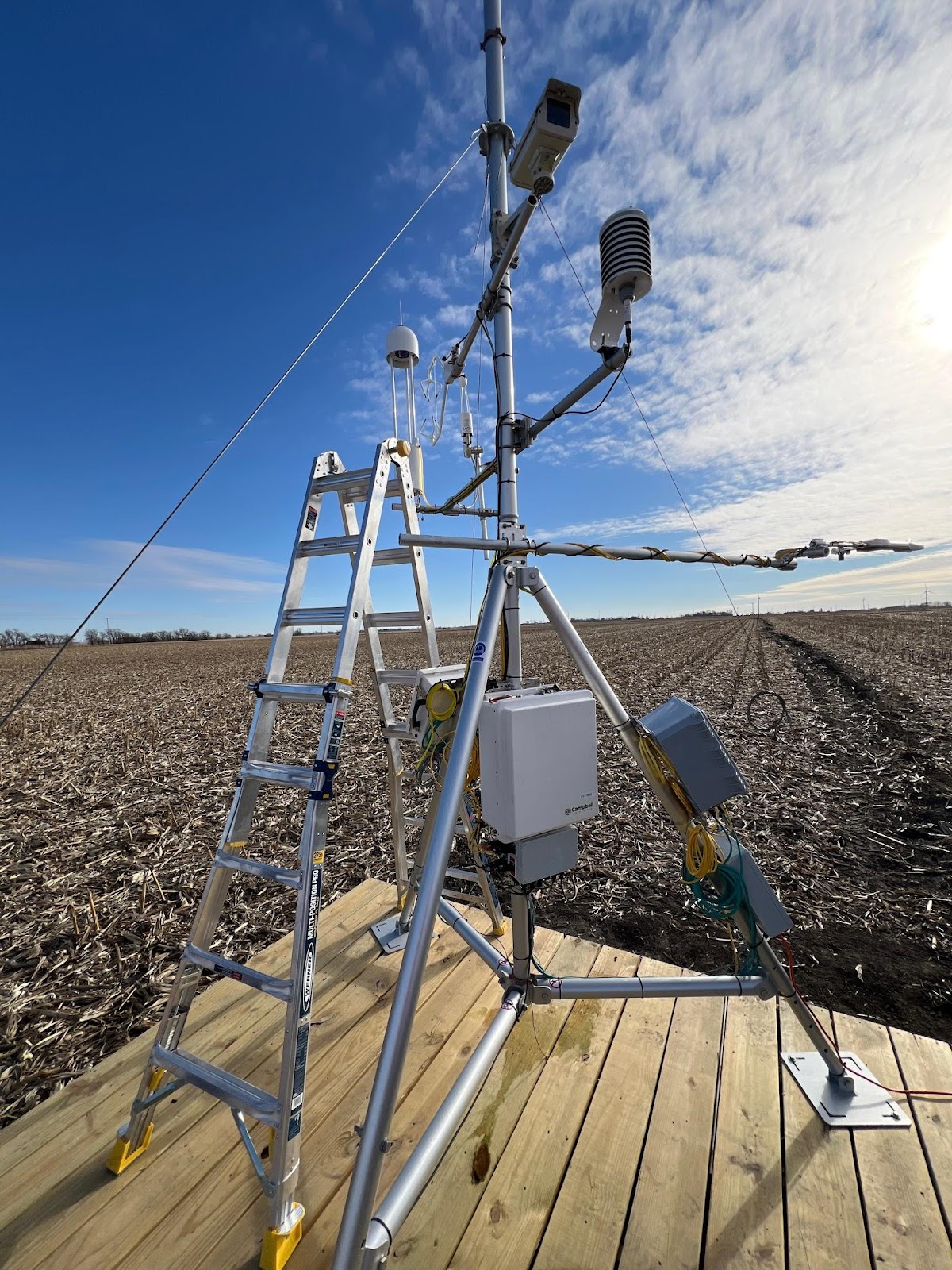 A science instrument is set up in an empty field. There is a step ladder to the side of the instrument. It is made of aluminum and has tripod legs.