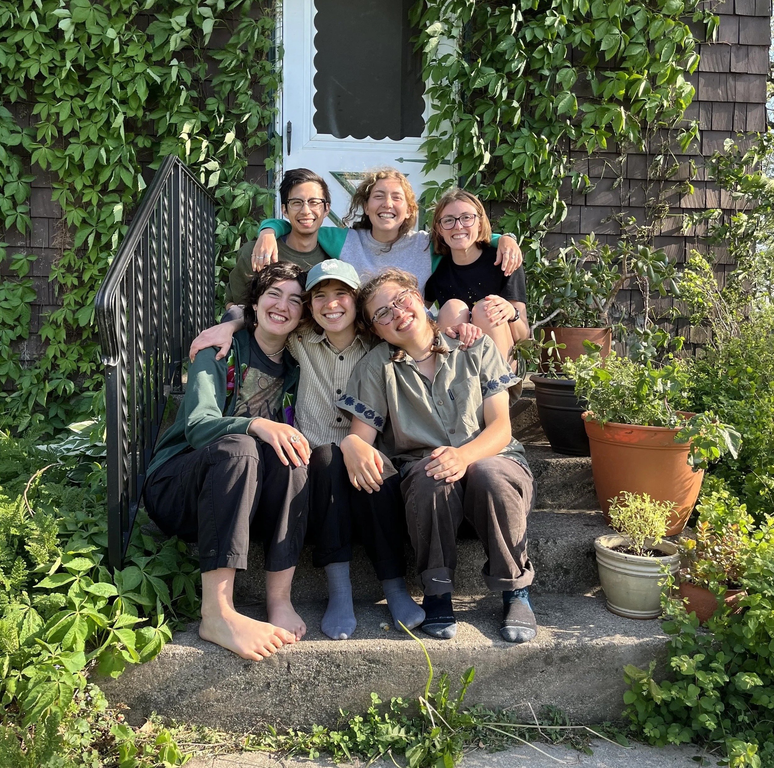 Six people sit on the steps in front of a ivy-covered house. They are all smiling and hugging each other. 